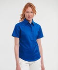 A person with red hair wearing a blue Russell J933F short-sleeve button-up shirt and white pants stands against a plain background.