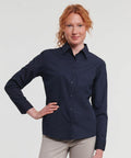 A person with red hair wearing a long-sleeve, dark blue button-up Russell J934F and light-colored pants poses with one hand on their hip against a plain background.