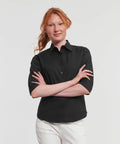 A person with long red hair is standing with arms crossed, wearing a black Russell J946F button-down shirt and white pants. The background is plain white.