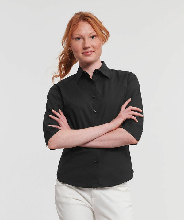 A person with long red hair is standing with arms crossed, wearing a black Russell J946F button-down shirt and white pants. The background is plain white.