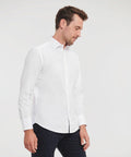 A man in a white long-sleeve J946M button-up shirt by Russell and dark pants stands against a plain background, looking down.