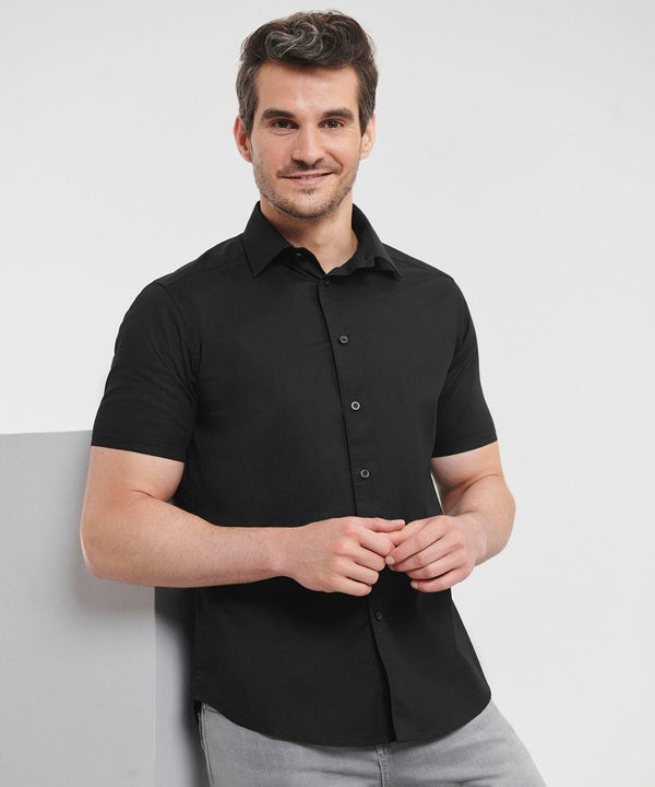 A man with short hair, wearing a black J947M shirt by Russell and gray pants, stands against a white background, smiling slightly.