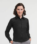 A person with short, curly dark hair wearing a black Russell J956F button-down shirt and gray pants stands against a plain white background, smiling with their hands in their pockets.