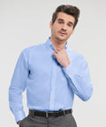 A man with dark hair wearing a light blue Russell J956M dress shirt and gray pants is adjusting his collar with his right hand.