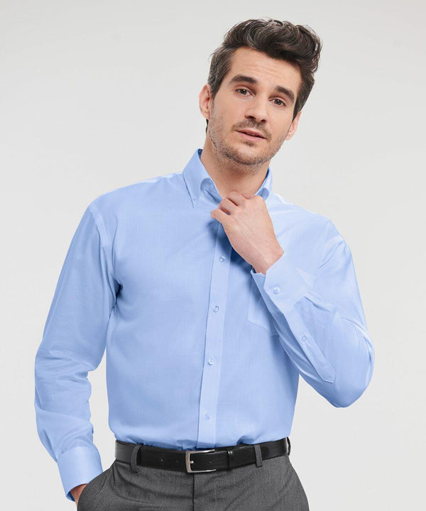 A man with dark hair wearing a light blue Russell J956M dress shirt and gray pants is adjusting his collar with his right hand.