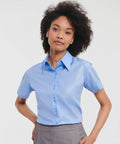 A person with curly hair is wearing a light blue, short-sleeved Russell J957F shirt and gray pants. They are standing in front of a white background.
