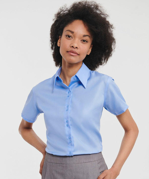 A person with curly hair is wearing a light blue, short-sleeved Russell J957F shirt and gray pants. They are standing in front of a white background.