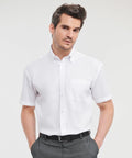A man with short brown hair wears a white short-sleeve button-up **Russell J957M** and gray trousers. He stands against a plain white background with his hands in his pockets.