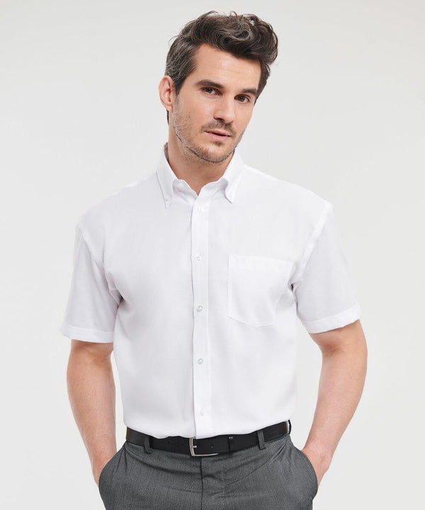 A man with short brown hair wears a white short-sleeve button-up **Russell J957M** and gray trousers. He stands against a plain white background with his hands in his pockets.