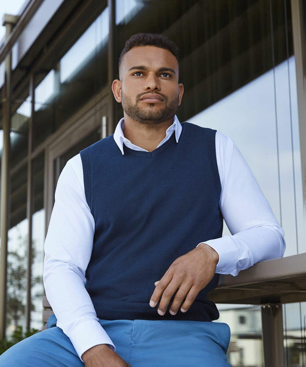 A man in a J962M by Russell, a white shirt and blue sweater vest, sits outside a modern building, looking into the distance.