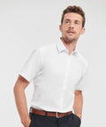 A man wearing a white short-sleeve button-up J963M shirt by Russell and dark pants with a brown belt stands against a plain background, looking upwards.