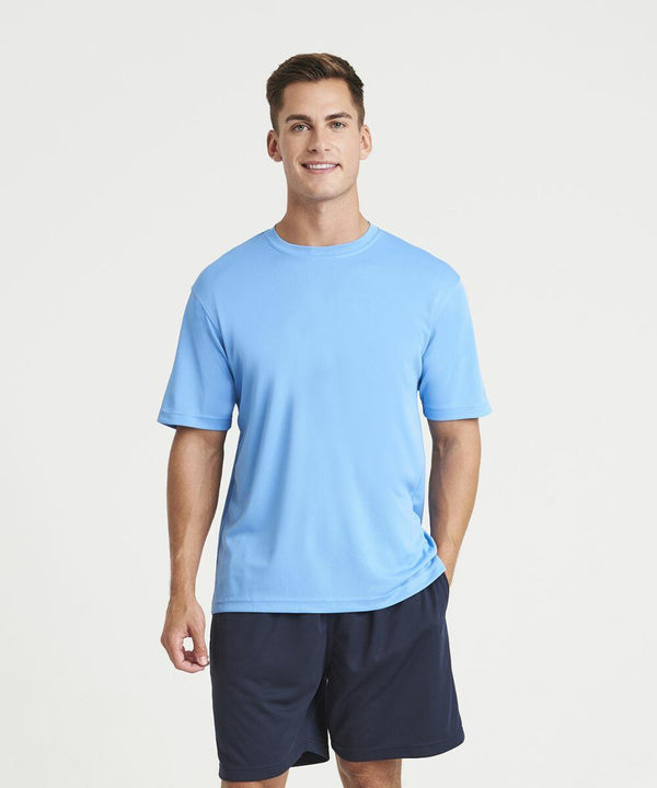 A person stands smiling, wearing a light blue AWDis Just Cool JC001 T-shirt and navy shorts. The background is plain white.