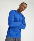 A person wearing a long-sleeved blue AWDis Just Cool JC002 shirt and gray pants stands against a plain background with their arms crossed.