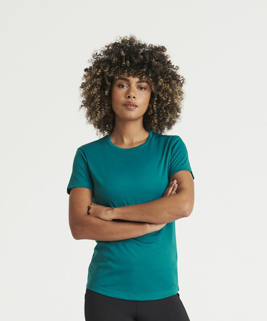 Person with curly hair wearing an AWDis Just Cool JC005 teal t-shirt and black pants, standing against a plain white background with arms crossed.