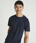 A person with short dark hair smiling and wearing an AWDis Just Cool JC011 plain dark blue T-shirt stands against a plain white background.