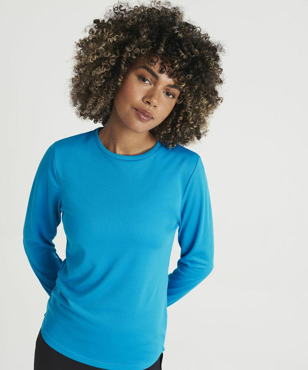 Person with curly hair wearing a long-sleeved blue AWDis Just Cool JC012, standing with hands behind their back, looking directly ahead against a plain white background.