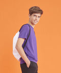 A young person with short hair wearing a purple shirt and black pants stands sideways against an orange background, carrying a white JC01J drawstring backpack by AWDis Just Cool.