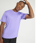 A person in a light purple AWDis Just Cool JC020 t-shirt and black pants is tilting their head to the side while touching their head with one hand.