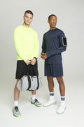 Two men in athletic wear stand against a plain background. One holds a duffel bag and wears a neon yellow JC023 shirt from AWDis Just Cool, while the other wears a dark shirt with an armband.
