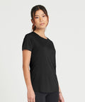 Person standing against a plain background wearing an AWDis Just Cool JC025 black short-sleeved shirt and dark pants, looking toward the camera.