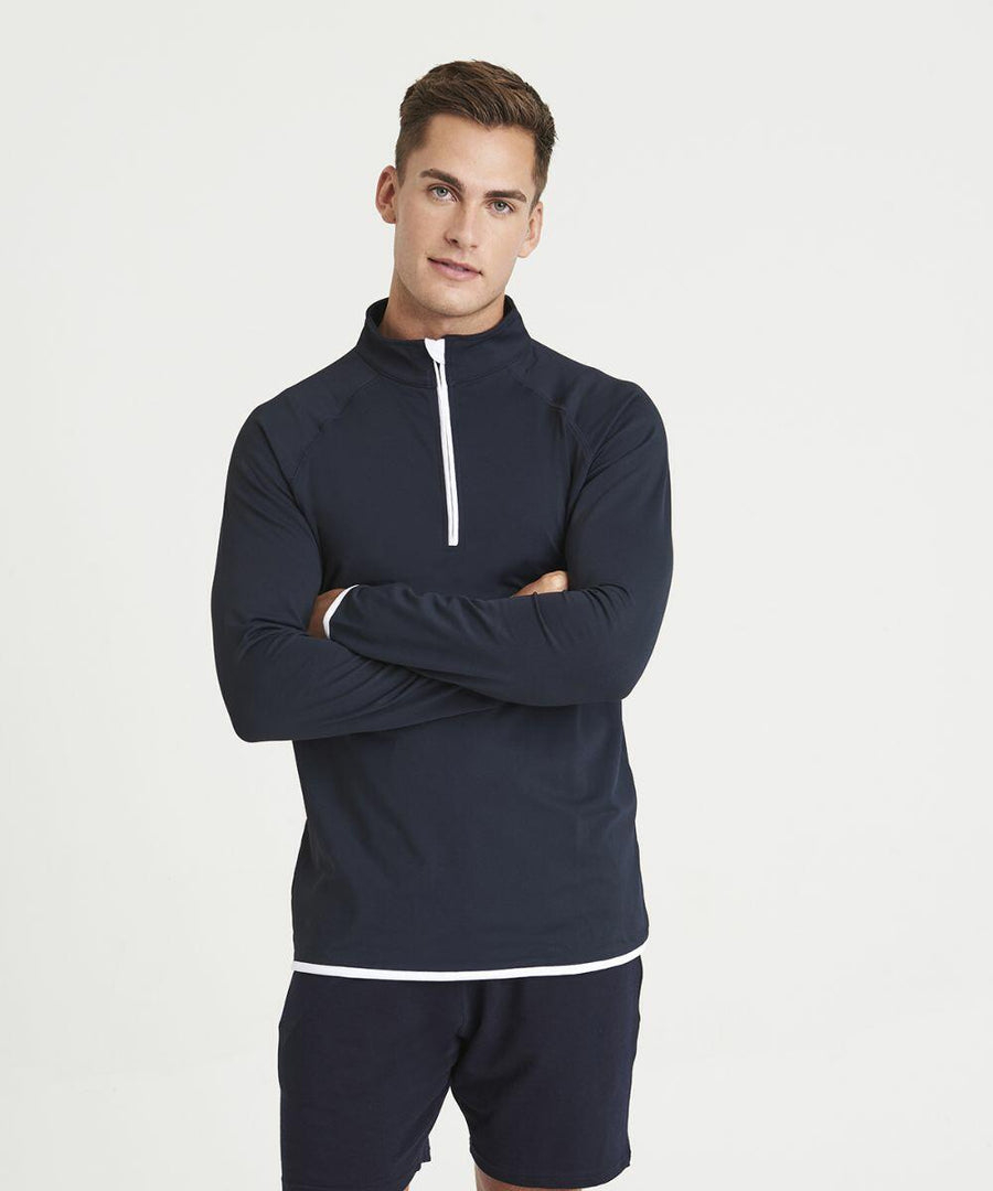 A man stands against a plain background, wearing an AWDis Just Cool JC031 navy long-sleeve half-zip pullover and navy shorts with his arms crossed.