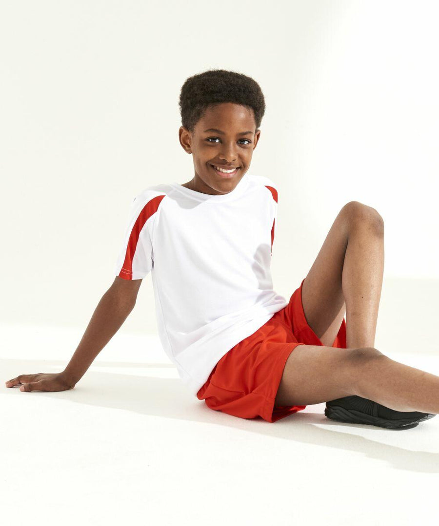 A young boy in a white JC03J shirt with red accents and red shorts from AWDis Just Cool is sitting on the floor smiling, one hand resting on the ground, and wearing black shoes.