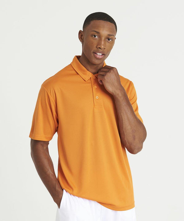 A man wearing an orange JC040 polo shirt from AWDis Just Cool and white pants stands against a plain white background, touching his collar with one hand and having his other hand in his pocket.