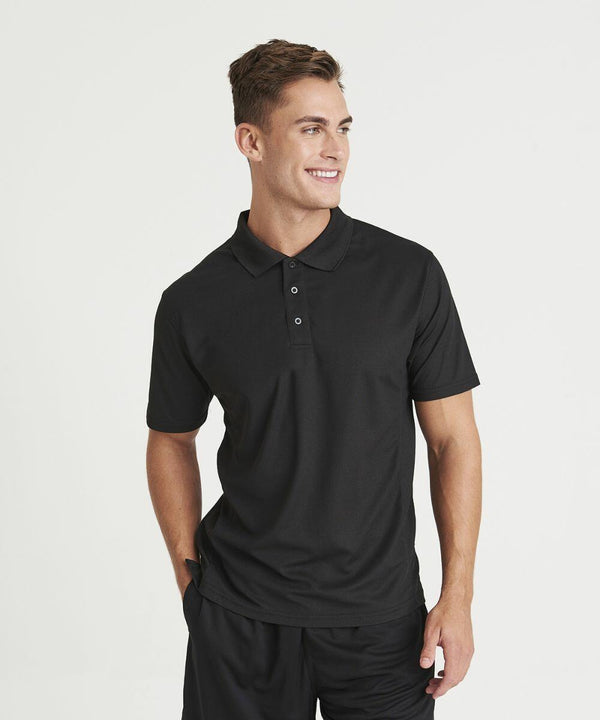 A person wearing a black AWDis Just Cool JC041 polo shirt and black shorts stands with one hand in their pocket, smiling slightly to the side against a plain white background.