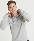 A person wearing a light gray JC052 hoodie by AWDis Just Cool looks at the camera while holding the hood strings with both hands.