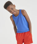 A boy wearing an AWDis Just Cool JC07J blue tank top and red shorts stands with his hands in his pockets, slightly leaning forward and smiling.