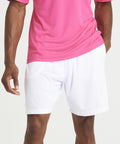 A person wearing a pink JC080 shirt from AWDis Just Cool and white shorts stands with their arms relaxed by their sides. The background is plain and white.