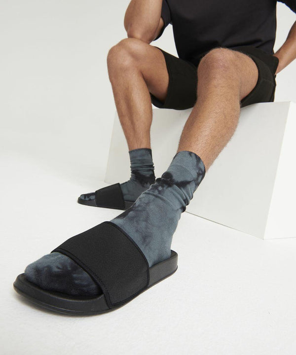 Person seated on a white bench wearing black shorts and a black JC092 t-shirt from AWDis Just Cool, with tie-dye socks and black slide sandals. Only the lower half of the body is shown.