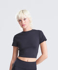 A woman with short, bleached blonde hair and a neutral expression models the AWDis Just Cool Women's recycled cropped T in black, paired with black bottoms against a plain light grey background.