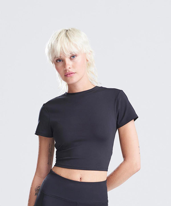 A woman with short, bleached blonde hair and a neutral expression models the AWDis Just Cool Women's recycled cropped T in black, paired with black bottoms against a plain light grey background.