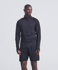 A man stands against a plain light background wearing the AWDis Just Cool Lightweight Active 1/4-zip in black and black shorts, looking slightly left with his left hand by his side and right hand near his thigh.