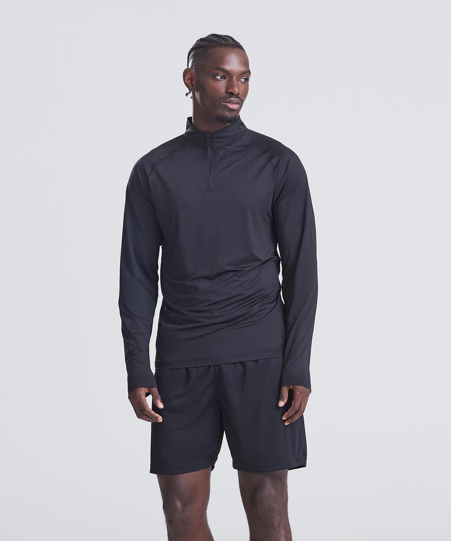 A man stands against a plain light background wearing the AWDis Just Cool Lightweight Active 1/4-zip in black and black shorts, looking slightly left with his left hand by his side and right hand near his thigh.