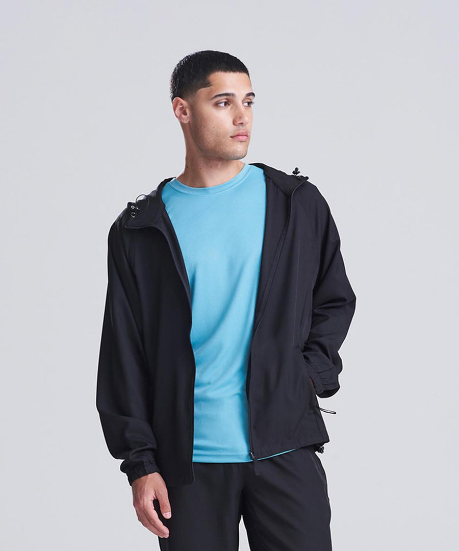 A man wears the AWDis Just Cool Active track jacket over a light blue T-shirt and black trousers, standing against a plain background with one hand in his pocket, looking to his left.
