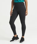 A person wearing black JC287 leggings, black sneakers, and a green top from AWDis Just Cool stands against a plain white background. The image shows them from the neck down.