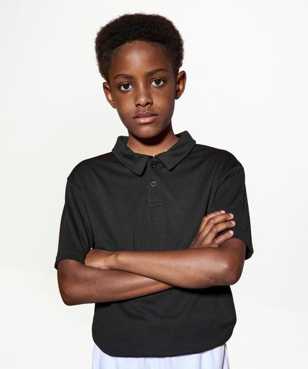 A young person stands with arms crossed, wearing an AWDis Just Cool JC40J black polo shirt and white pants, against a plain white background.