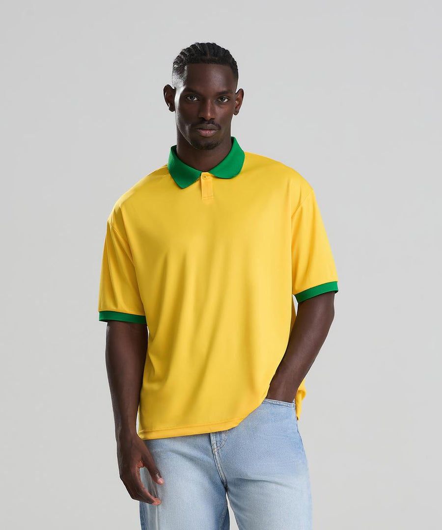A man with a neutral expression, one hand in his pocket, wears the AWDis Just Cool Retro sports jersey—yellow with green collar and sleeve trim—and light blue jeans against a plain background.