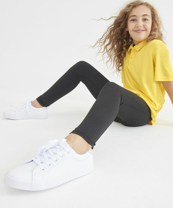 A young girl with long curly hair, wearing an AWDis Just Cool JC87J yellow shirt, black leggings, and white sneakers, sits on the floor against a plain white background.