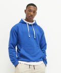 A person stands against a white background, wearing an AWDis Just Hoods JH003 blue hoodie with white drawstrings and beige pants.