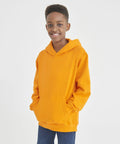Young person in an AWDis Just Hoods JH01J orange hoodie and blue jeans smiles while standing with hands in front pocket against a plain white background.