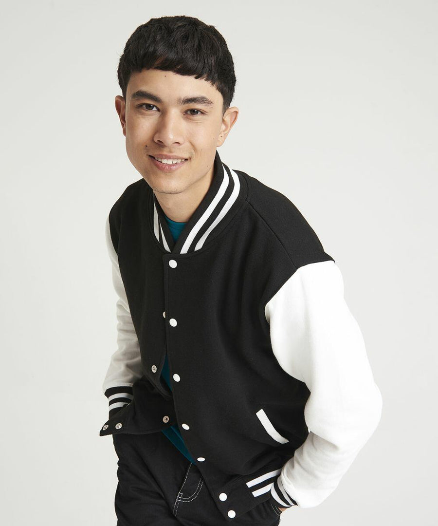 Person smiling and wearing an AWDis Just Hoods JH043 black and white varsity jacket over a blue shirt, leaning slightly forward against a plain white background.