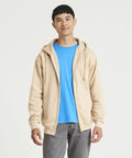 A man wearing an AWDis Just Hoods JH050 beige zip-up hoodie over a blue T-shirt and gray pants stands against a plain white background.