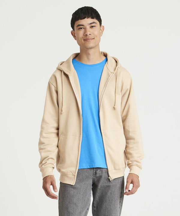 A man wearing an AWDis Just Hoods JH050 beige zip-up hoodie over a blue T-shirt and gray pants stands against a plain white background.