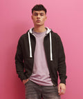 A person with short dark hair, dressed in an AWDis Just Hoods JH052 with white drawstrings and a light purple shirt, stands against a pink wall with hands in pockets.