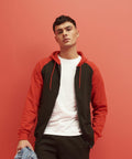 A person stands against a red background wearing a black and red JH063 hoodie from AWDis Just Hoods, a white shirt, and holding a pair of jeans.