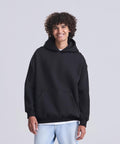 A person with curly hair is standing and smiling, wearing the Vision heavyweight hoodie from AWDis Just Hoods over a white shirt and light blue jeans, against a plain light grey background.