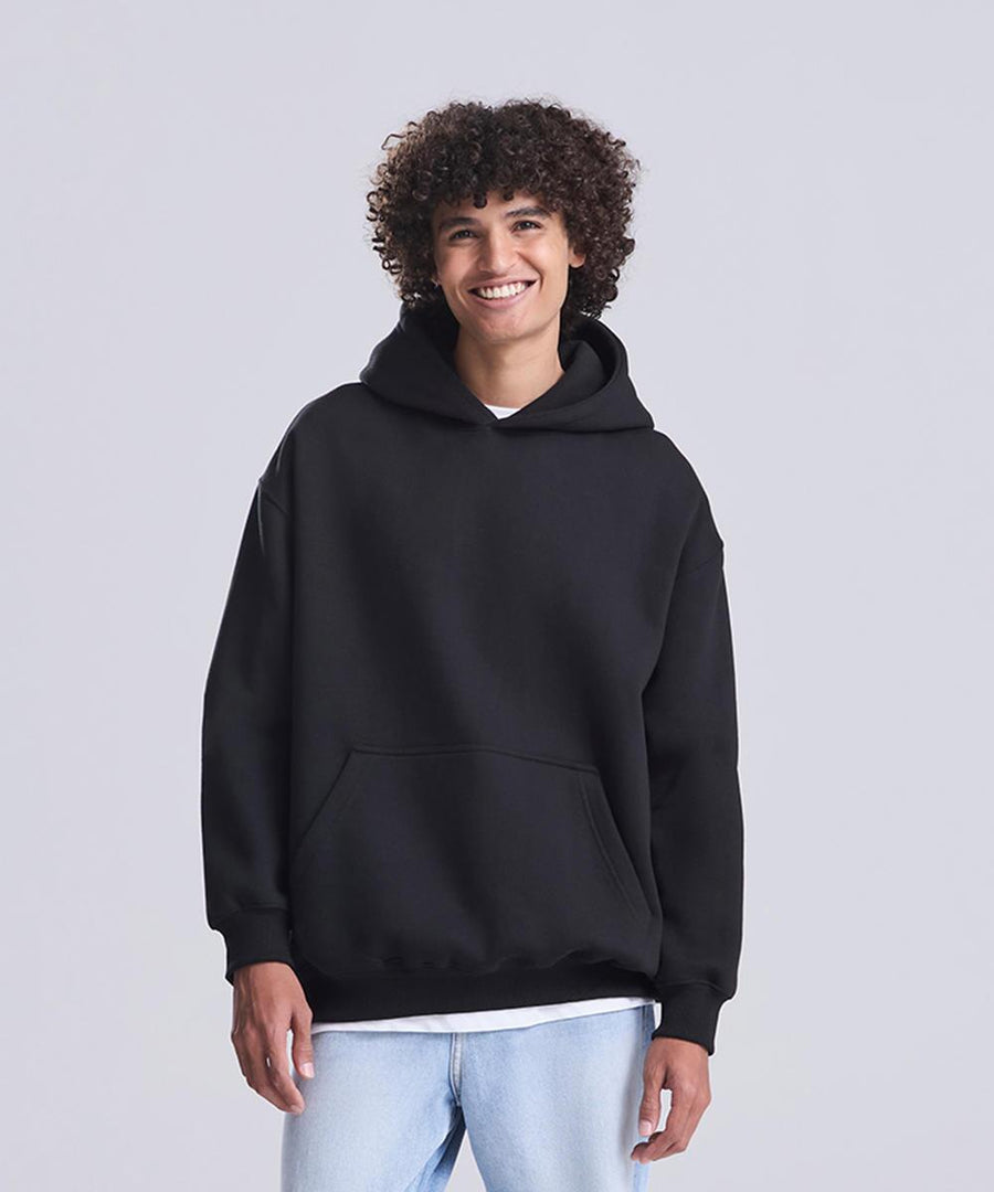 A person with curly hair is standing and smiling, wearing the Vision heavyweight hoodie from AWDis Just Hoods over a white shirt and light blue jeans, against a plain light grey background.
